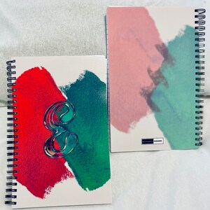 Op de afbeelding: Twee spiraalgebonden notitieboekjes met abstracte aquarelontwerpen. Een notitieboekje heeft een rood en groen ontwerp met een doorzichtig plastic nummer "8" op de voorkant. Het andere notitieboekje heeft een roze en groen ontwerp met de tekst "DYNAMIC DESIGNS" op de voorkant.