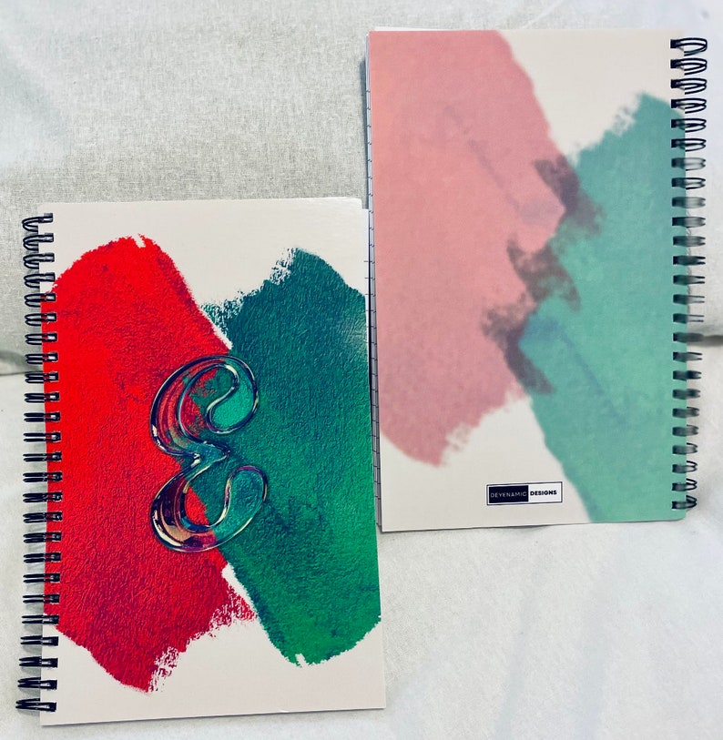 Op de afbeelding: Twee spiraalgebonden notitieboekjes met abstracte aquarelontwerpen. Een notitieboekje heeft een rood en groen ontwerp met een doorzichtig plastic nummer "8" op de voorkant. Het andere notitieboekje heeft een roze en groen ontwerp met de tekst "DYNAMIC DESIGNS" op de voorkant.