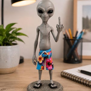 May include: A gray alien figurine standing on a circular stone-textured base. The figure has large black eyes, a small smile, and is wearing blue board shorts with a vibrant pink and yellow hibiscus floral pattern, white drawstrings, and black flip-flops.