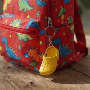 May include: A bright red backpack featuring a repeating pattern of colourful dinosaurs and yellow lightning bolts. A small, yellow, perforated clog-shaped keychain is attached to the front zip pull of the backpack with a silver metal ring.