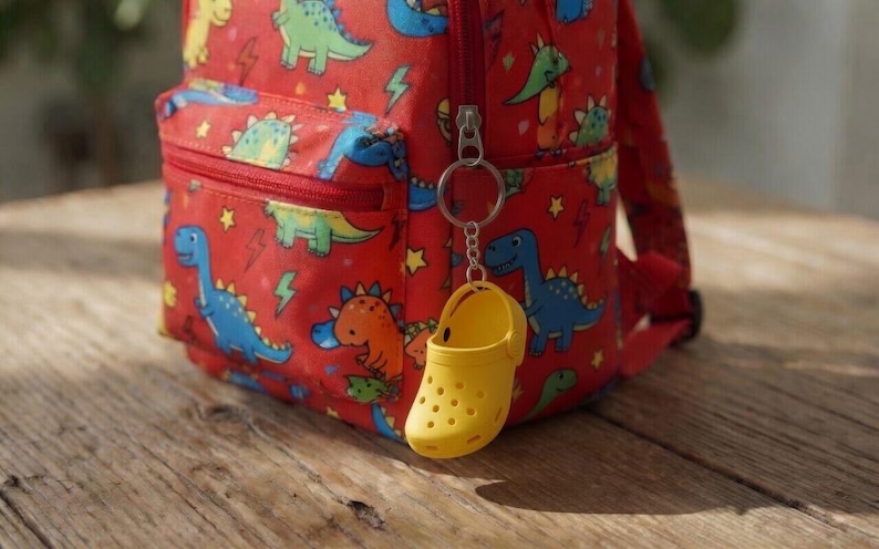 May include: A bright red backpack featuring a repeating pattern of colourful dinosaurs and yellow lightning bolts. A small, yellow, perforated clog-shaped keychain is attached to the front zip pull of the backpack with a silver metal ring.