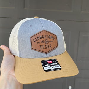 Puede incluir: Gorra de camionero gris, beige y blanca con un parche de cuero marrón que dice "Georgetown Texas" en forma de hexágono. La gorra tiene una etiqueta Richardson 115.