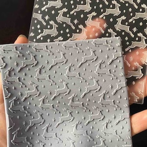 May include: A clear plastic sheet with a repeating pattern of white reindeer and snowflakes. The sheet is placed on top of a grey silicone sheet with the same pattern embossed into it.