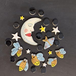 May include: A collection of ceramic mosaic tiles featuring a crescent moon with a face, four fireflies with blue wings and yellow bodies, a moth, several white and yellow stars, and numerous small black circular tiles arranged in a circular pattern.