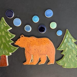 May include: A set of ceramic pieces featuring a brown bear, two green pine trees with textured needles, and seven small circular discs in shades of blue, light blue, and pale green, all arranged on a dark surface to create a forest scene.