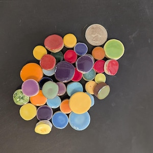 May include: A collection of colorful, ceramic, circular tiles in various sizes and hues, including red, yellow, orange, purple, and green. A U.S. quarter coin is included for size reference. The tiles are arranged on a dark surface.