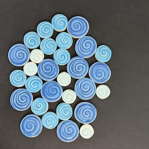 May include: A collection of circular ceramic tiles in various shades of blue and light blue, each featuring a raised spiral pattern on the surface. The tiles vary in size and are arranged in a cluster, displaying glossy finishes and smooth edges.