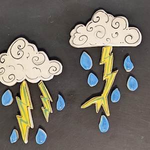 May include: Two ceramic cloud and lightning bolt decorations. The clouds are white with black outlines and swirling details. Yellow lightning bolts with blue accents and blue teardrop-shaped rain complete the weather-themed design.