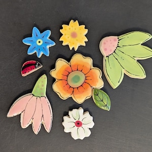 May include: A collection of ceramic decorative pieces including a blue star-shaped flower, a yellow flower, a large orange flower with a green leaf, a pink drooping flower, a white flower with a red center, a green leaf cluster, and a red ladybug.