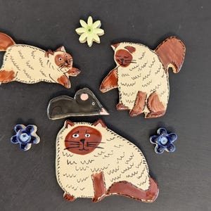 May include: A set of ceramic decorative pieces featuring three cream and brown cats, one small black mouse, one pale yellow flower, and two small blue flowers. The cats have dark brown faces, ears, and tails with cream-colored bodies.
