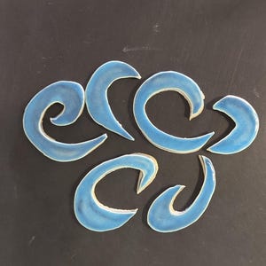 May include: A collection of seven light blue ceramic pieces, each with a curved, comma-like shape. The pieces are arranged on a dark gray surface. The ceramic has a glossy finish.