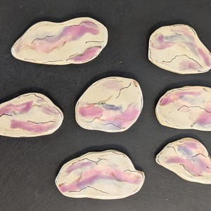 May include: Seven irregular, flat ceramic pieces with a cream-colored base and abstract washes of purple and blue. Each piece features thin, dark, hand-drawn lines that mimic natural stone veining, creating a unique pattern on every individual item.