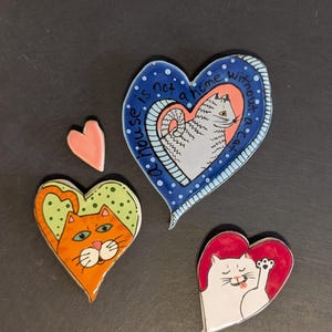 May include: Three heart-shaped ceramic ornaments and one small pink heart. One blue heart features a gray cat illustration with the text A house is not a home without a cat. One orange heart shows a cat face, and one red heart shows a white cat.