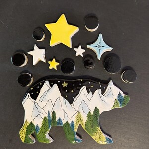 May include: A ceramic bear-shaped wall plaque featuring a painted mountain landscape with green pine trees and a starry night sky, accompanied by several separate ceramic star and circle shapes in yellow, white, light blue, and black colors.