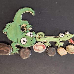 May include: A whimsical illustration featuring a green lizard and a smaller lizard with a red ladybug. The lizards are positioned above a row of painted stones. The artwork is on a dark background.