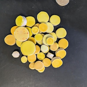 May include: A collection of yellow and orange ceramic tiles in various sizes and shades. The tiles are round, with some having a white edge. The arrangement includes tiles with speckled and solid surfaces, creating a visually appealing assortment.