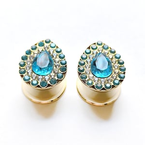 Può includere: Coppia di dilatatori per orecchie dorati con una gemma blu a forma di lacrima circondata da strass trasparenti e blu.