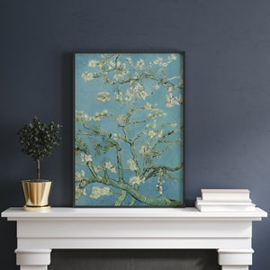 Puede incluir: Una impresi&oacute;n enmarcada de una pintura que representa un almendro en flor blanco sobre un fondo de cielo azul. La pintura est&aacute; en un marco negro y est&aacute; colgada en una repisa de chimenea blanca.