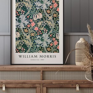 May include: A framed print of a vintage floral pattern by William Morris, titled "Golden Lily" and dated 1880. The design features a repeating pattern of stylised lilies, leaves, and flowers in shades of green, blue, pink, and yellow.