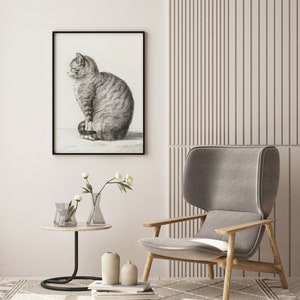 May include: A black and white drawing of a cat sitting on a white background. The drawing is framed in a black frame and is hanging on a white wall. A gray armchair with wooden legs is in the foreground. There are two clear glass vases with white flowers on a black side table.