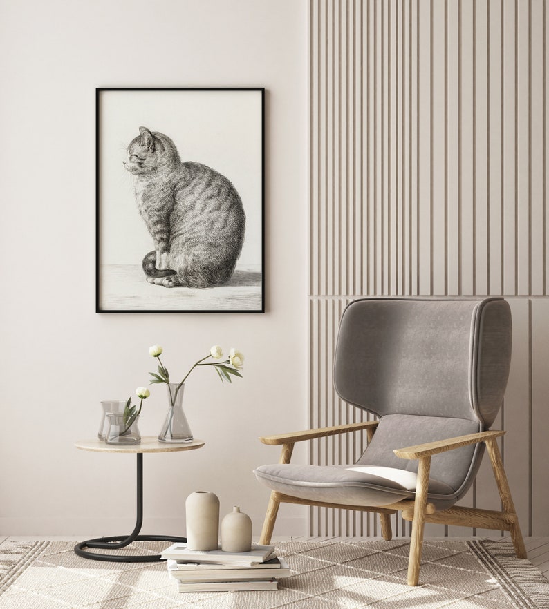 May include: A black and white drawing of a cat sitting on a white background. The drawing is framed in a black frame and is hanging on a white wall. A gray armchair with wooden legs is in the foreground. There are two clear glass vases with white flowers on a black side table.