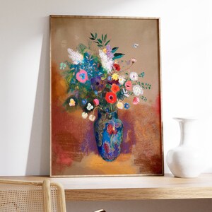 May include: A framed art print featuring a vibrant floral still life. The artwork showcases a blue vase filled with colorful flowers, including red, pink, and white blooms. The background has warm tones of brown and red. The frame is a light brown color.