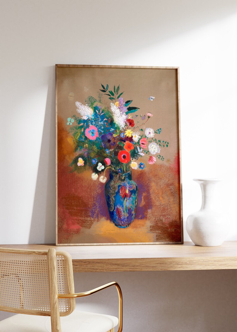 May include: A framed art print featuring a vibrant floral still life. The artwork showcases a blue vase filled with colorful flowers, including red, pink, and white blooms. The background has warm tones of brown and red. The frame is a light brown color.