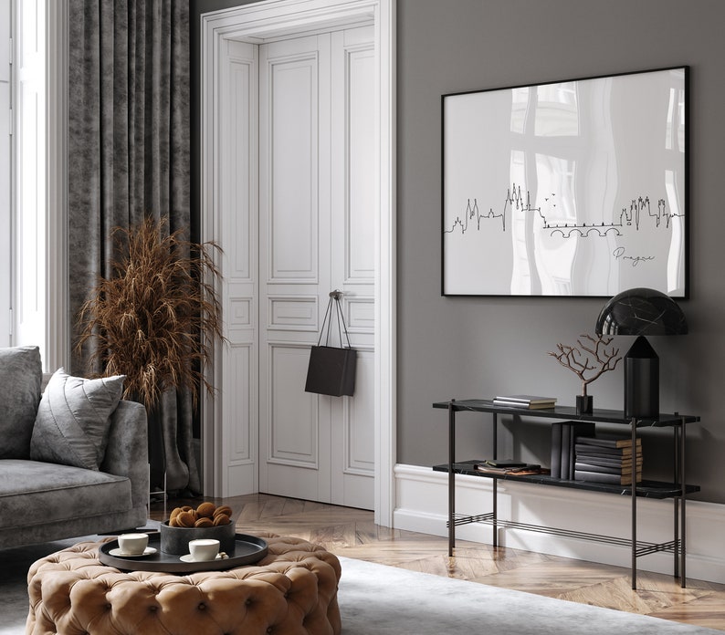 May include: A stylish living room featuring a grey sofa, a buttoned ottoman, and a framed print of a city skyline. A black handbag hangs on the white door. A black marble lamp and console table add to the decor.