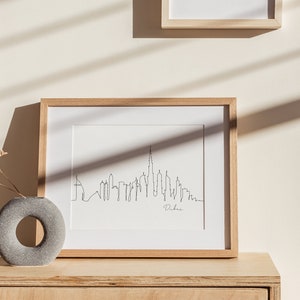 May include: A framed black and white line drawing of the Dubai skyline with the word "Dubai" written below the skyline. The frame is made of light wood and is sitting on a light wood surface.