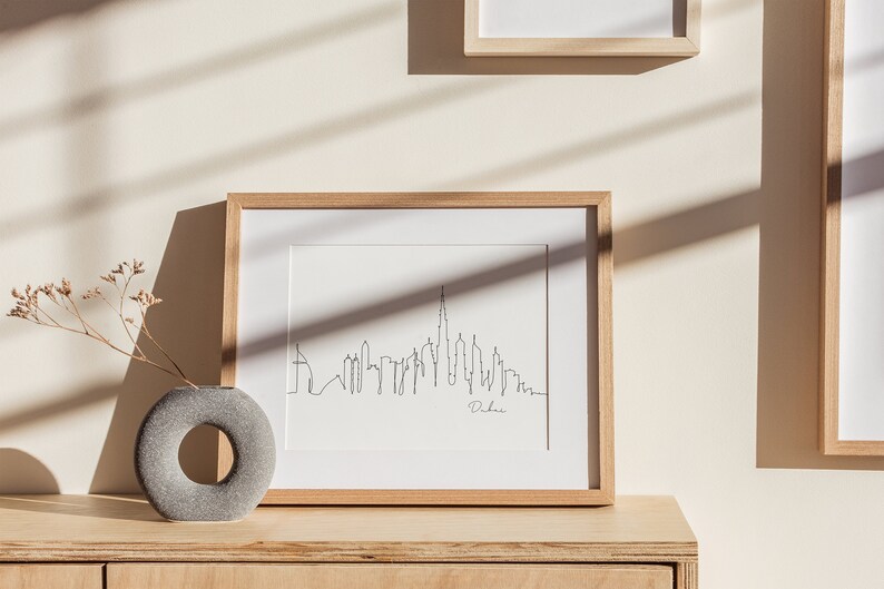 May include: A framed black and white line drawing of the Dubai skyline with the word "Dubai" written below the skyline. The frame is made of light wood and is sitting on a light wood surface.