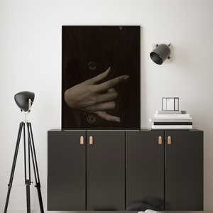 Op de afbeelding: Een zwart-witfoto van een hand die een gebaar maakt, ingelijst in een zwart frame en hangend aan een witte muur boven een zwarte kast met twee deuren. De kast heeft twee bruine lederen handgrepen.