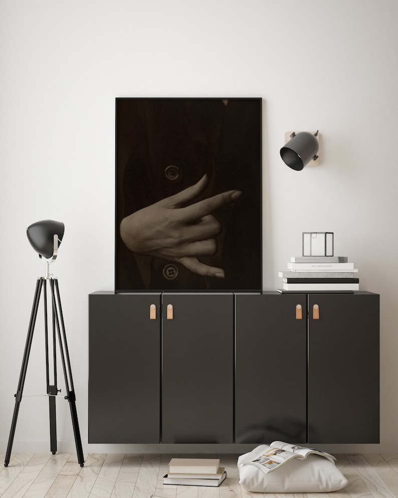 Op de afbeelding: Een zwart-witfoto van een hand die een gebaar maakt, ingelijst in een zwart frame en hangend aan een witte muur boven een zwarte kast met twee deuren. De kast heeft twee bruine lederen handgrepen.