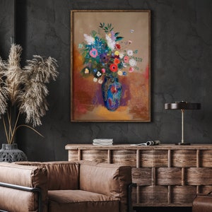 May include: A framed floral painting featuring a vibrant bouquet in a blue vase. The artwork showcases a mix of colorful flowers, including red, pink, and white blooms, set against a warm, textured background. The painting is displayed in a room with a dark wall and wooden furniture.