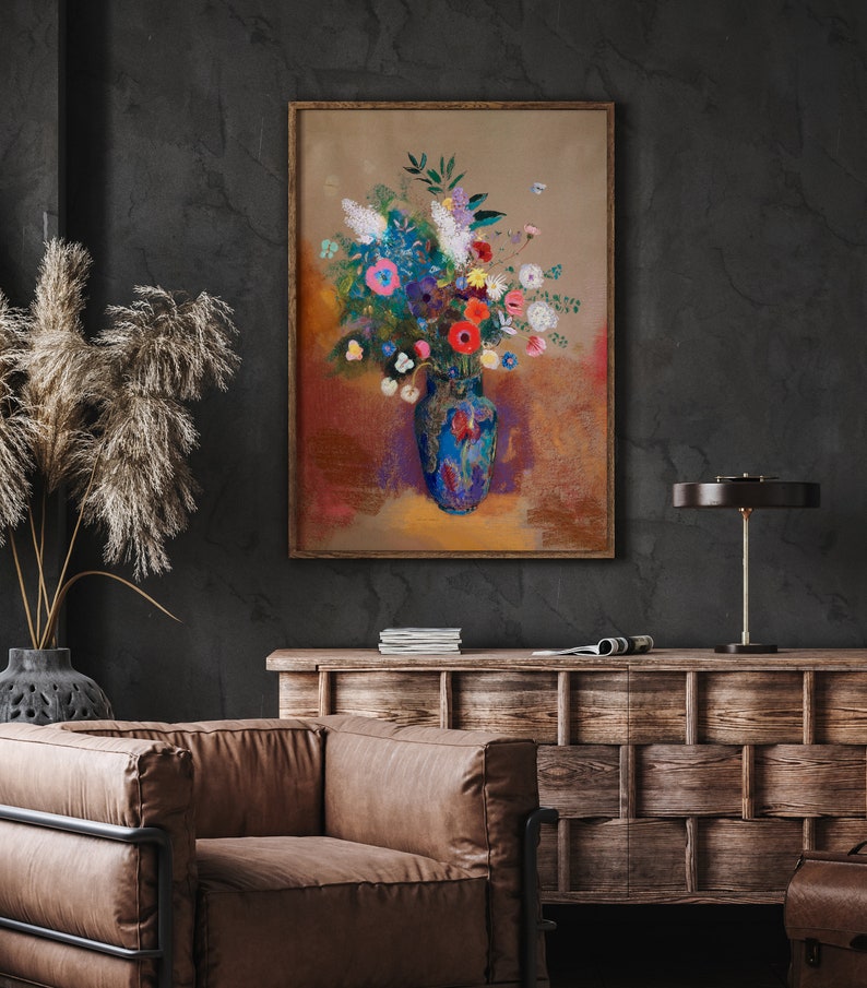 May include: A framed floral painting featuring a vibrant bouquet in a blue vase. The artwork showcases a mix of colorful flowers, including red, pink, and white blooms, set against a warm, textured background. The painting is displayed in a room with a dark wall and wooden furniture.