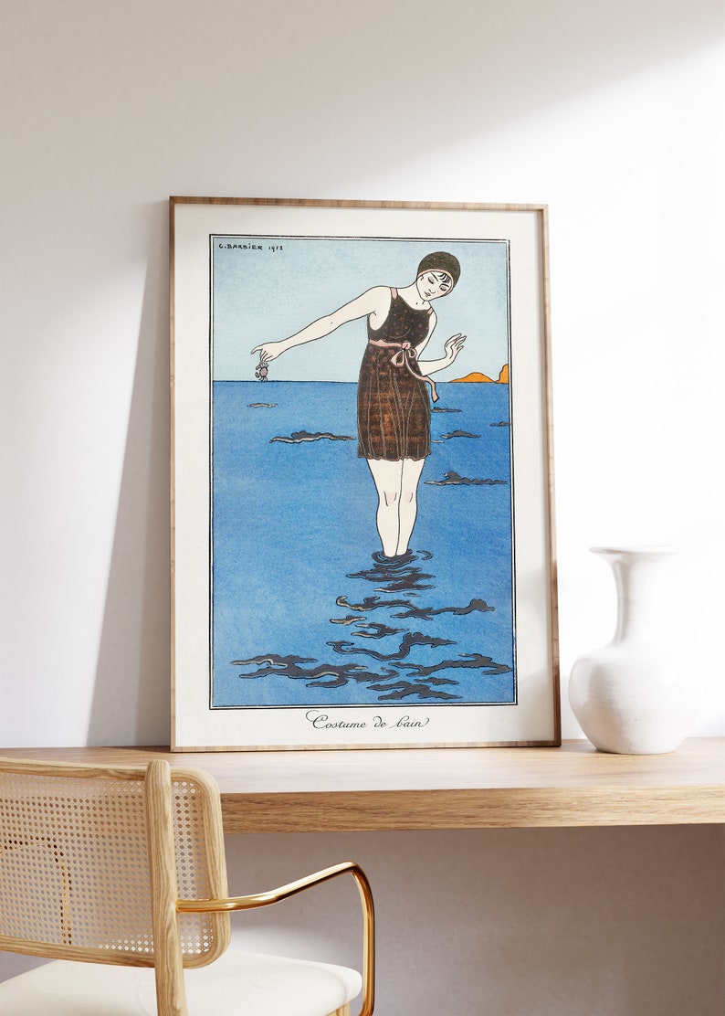 May include: A vintage illustration of a woman in a black swimsuit standing in the ocean. The illustration is in a frame and the text "Costume de bain" is at the bottom of the image.