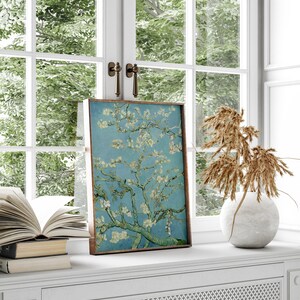 Puede incluir: Una impresi&oacute;n enmarcada de una pintura que representa un almendro en flor con flores blancas sobre un fondo de cielo azul. La impresi&oacute;n se muestra en un estante blanco con un jarr&oacute;n de flores secas y una pila de libros.