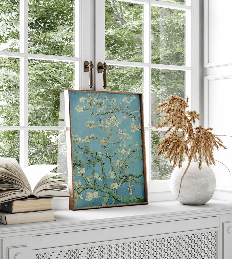 Puede incluir: Una impresi&oacute;n enmarcada de una pintura que representa un almendro en flor con flores blancas sobre un fondo de cielo azul. La impresi&oacute;n se muestra en un estante blanco con un jarr&oacute;n de flores secas y una pila de libros.
