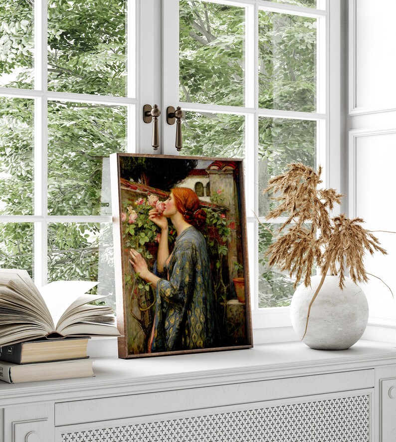 Op de afbeelding: Een ingelijst schilderij van een vrouw met rood haar die aan een roze roos ruikt in een tuin. De vrouw draagt een blauwe en gouden gepatroneerde robe. Het schilderij is geplaatst tegen een witte muur met een raam erachter.