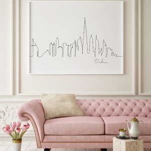 May include: A black and white line drawing of the Dubai skyline with the text "Dubai" below the drawing. The drawing is framed and hanging on a white wall above a pink sofa.