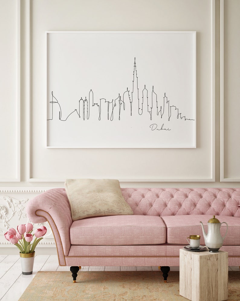 May include: A black and white line drawing of the Dubai skyline with the text "Dubai" below the drawing. The drawing is framed and hanging on a white wall above a pink sofa.