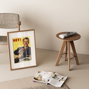 May include: A framed poster with the word "HET!" and a man in a suit holding up a glass of liquid. The poster is leaning against a light brown chair with a gray fabric seat. A small round wooden table with three legs sits in the foreground with a rolled up newspaper on top.