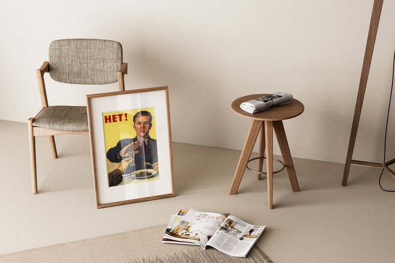 May include: A framed poster with the word "HET!" and a man in a suit holding up a glass of liquid. The poster is leaning against a light brown chair with a gray fabric seat. A small round wooden table with three legs sits in the foreground with a rolled up newspaper on top.