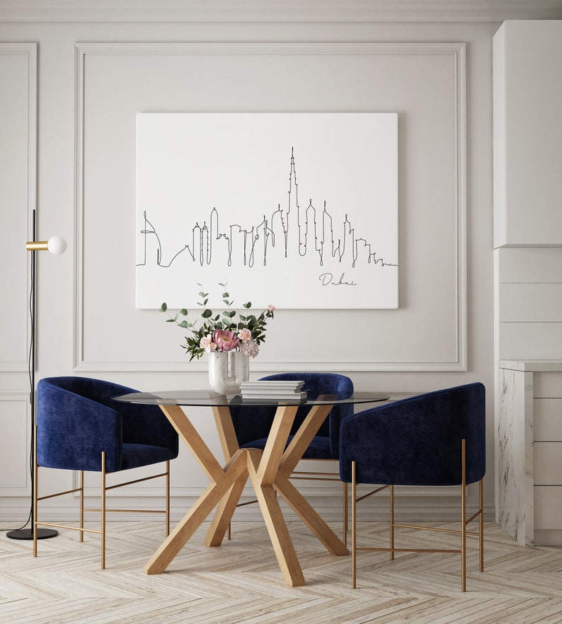 May include: A dining room with a glass-top table and four blue velvet chairs. The table has a wooden base with a unique X-shaped design. A black floor lamp with a white globe is in the corner of the room. A framed print of a line drawing of the Dubai skyline hangs on the wall.