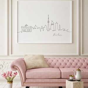 Puede incluir: Un sof&aacute; rosa capiton&eacute; con un coj&iacute;n blanco y un dibujo en blanco y negro enmarcado del horizonte de Berl&iacute;n. El dibujo dice "Berl&iacute;n" en la parte inferior. El sof&aacute; est&aacute; frente a una pared blanca con moldura blanca.