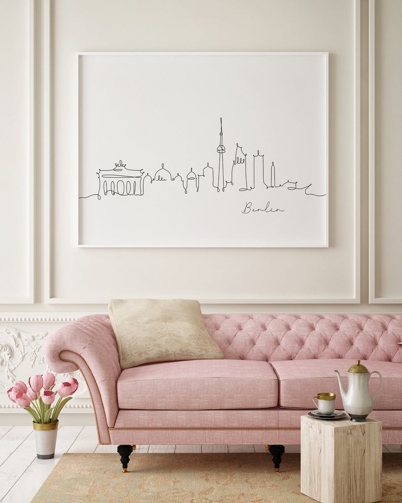 Puede incluir: Un sof&aacute; rosa capiton&eacute; con un coj&iacute;n blanco y un dibujo en blanco y negro enmarcado del horizonte de Berl&iacute;n. El dibujo dice "Berl&iacute;n" en la parte inferior. El sof&aacute; est&aacute; frente a una pared blanca con moldura blanca.