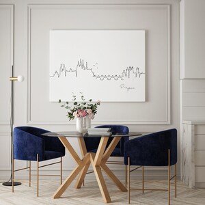May include: A contemporary dining set with a round glass table, light wood base, and four navy blue velvet chairs with gold-coloured legs. A white canvas displays a black line drawing of the Prague skyline. A vase of flowers sits on the table, beneath the artwork.