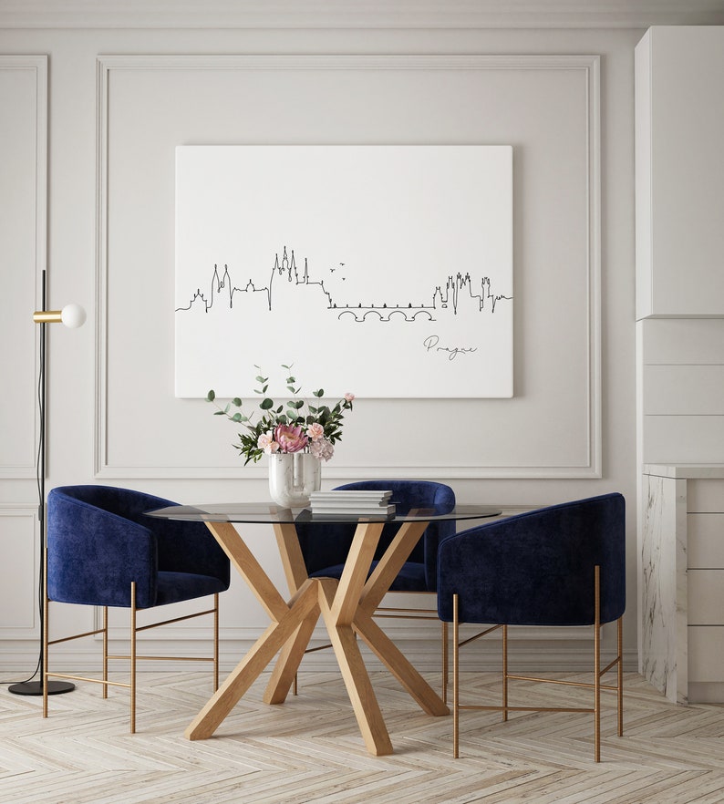 May include: A contemporary dining set with a round glass table, light wood base, and four navy blue velvet chairs with gold-coloured legs. A white canvas displays a black line drawing of the Prague skyline. A vase of flowers sits on the table, beneath the artwork.