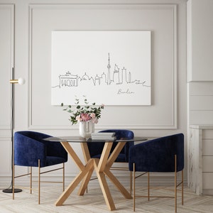 Puede incluir: Un comedor con una mesa redonda de vidrio y cuatro sillas de terciopelo azul. La mesa tiene una base de madera con un dise&ntilde;o en X &uacute;nico. Una l&aacute;mpara de pie negra con un globo blanco est&aacute; en la esquina de la habitaci&oacute;n. Una impresi&oacute;n enmarcada de un dibujo lineal del horizonte de Berl&iacute;n cuelga en la pared sobre la mesa. La impresi&oacute;n incluye la palabra "Berl&iacute;n" debajo del dibujo.