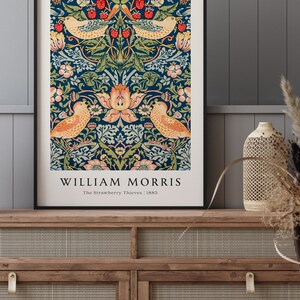 Op de afbeelding: Een ingelijste print van een vintage ontwerp van William Morris met een blauwe achtergrond met een bloemenpatroon en twee vogels. De tekst "William Morris" staat onderaan de print, samen met de titel "The Strawberry Thieves" en het jaar "1883".