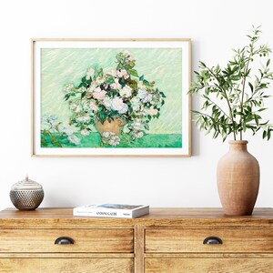 K&ouml;nnte beinhalten: Ein gerahmtes Gem&auml;lde mit einer Vase voller wei&szlig;er Rosen. Das Gem&auml;lde ist in einem hellen Holzrahmen gefasst und h&auml;ngt an einer wei&szlig;en Wand. Die Wand ist au&szlig;erdem mit einer Vase mit gr&uuml;nen Bl&auml;ttern und einer kleinen dekorativen Schale verziert.